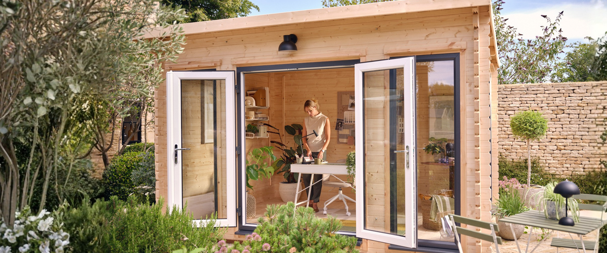 A woman working at a desk inside a Waltons wooden garden office, with white double doors open onto a sunny patio garden